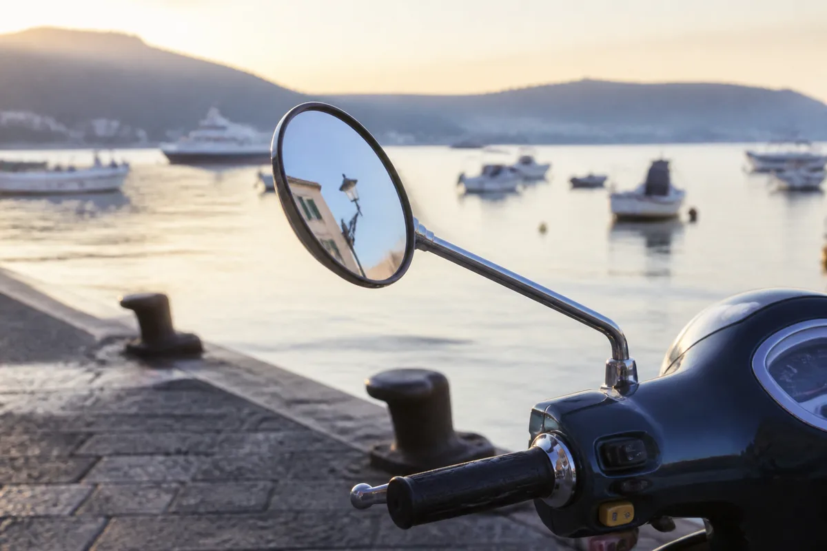 Scooter garé le long d&rsquo;un quai en bord de mer au petit matin, guidon et rétroviseur au premier plan